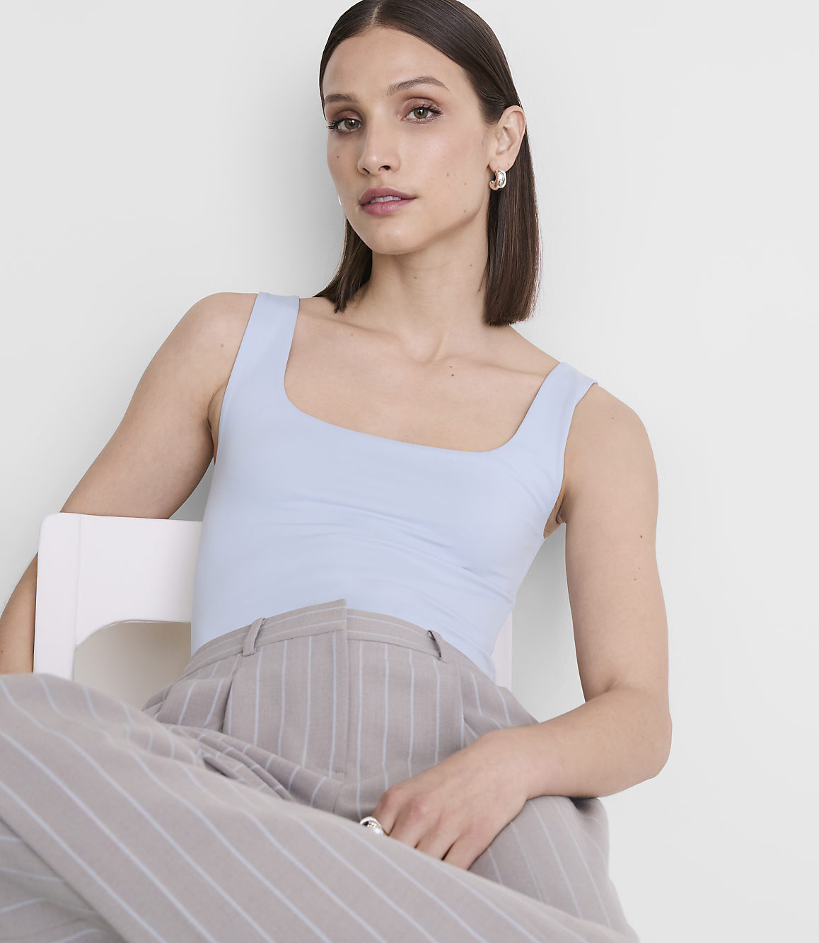 A woman in a light blue tank top and pinstripe pants sits on a white chair against a plain white background.