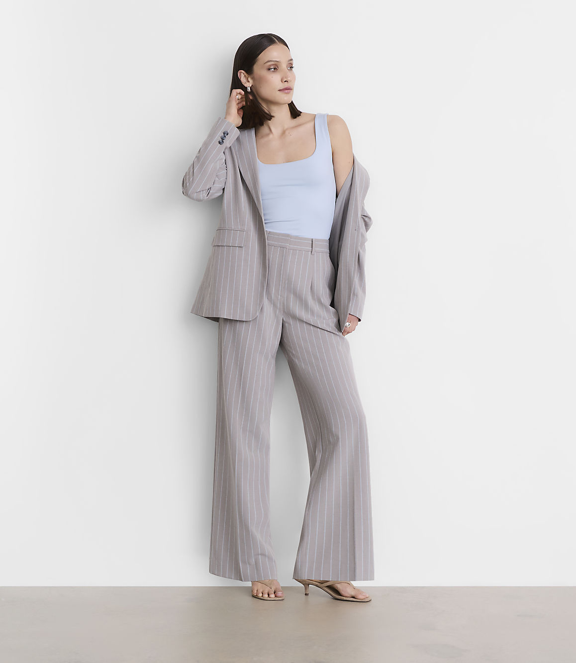 A woman in a light blue tank top and a pinstripe suit stands against a plain white wall.
