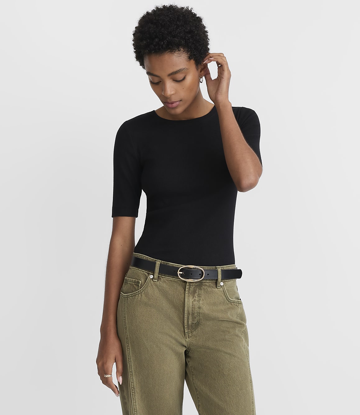 A woman in a black top and olive green pants stands against a plain white background.