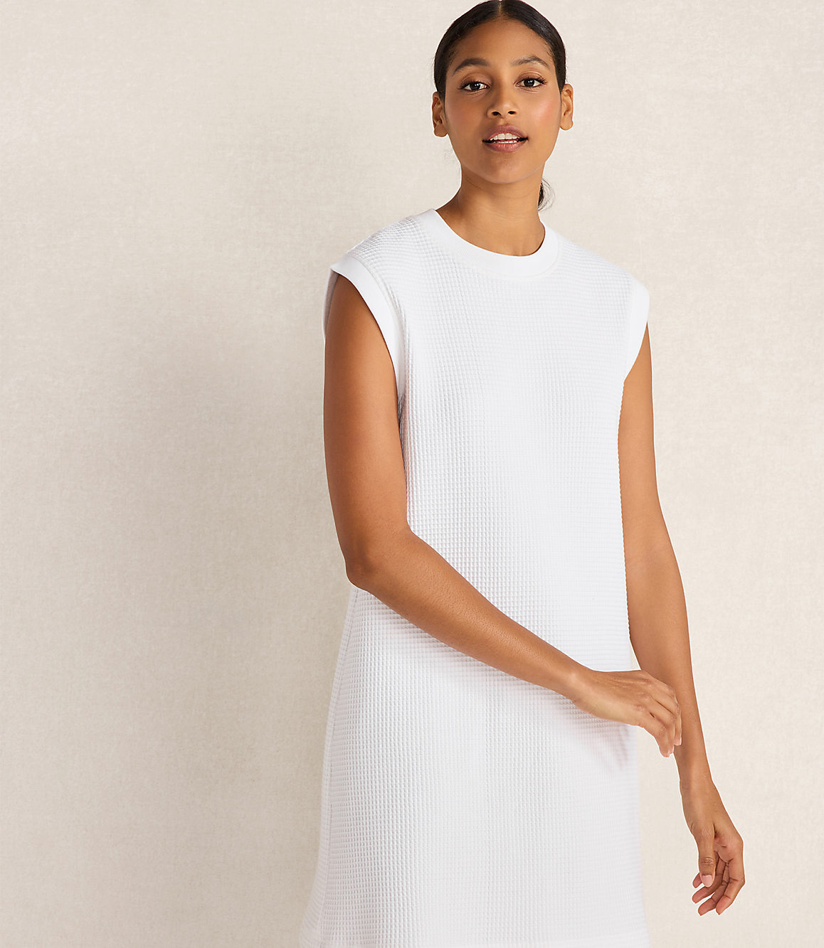 A woman in a white waffle-knit dress stands against a textured, light-colored wall.
