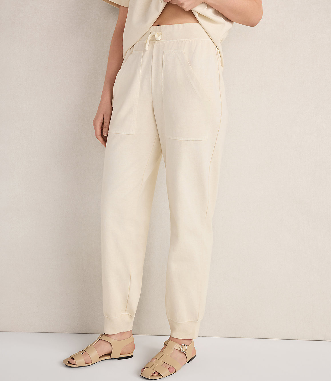 A person wearing cream-colored sweatpants and sandals stands against a textured, light-colored wall.