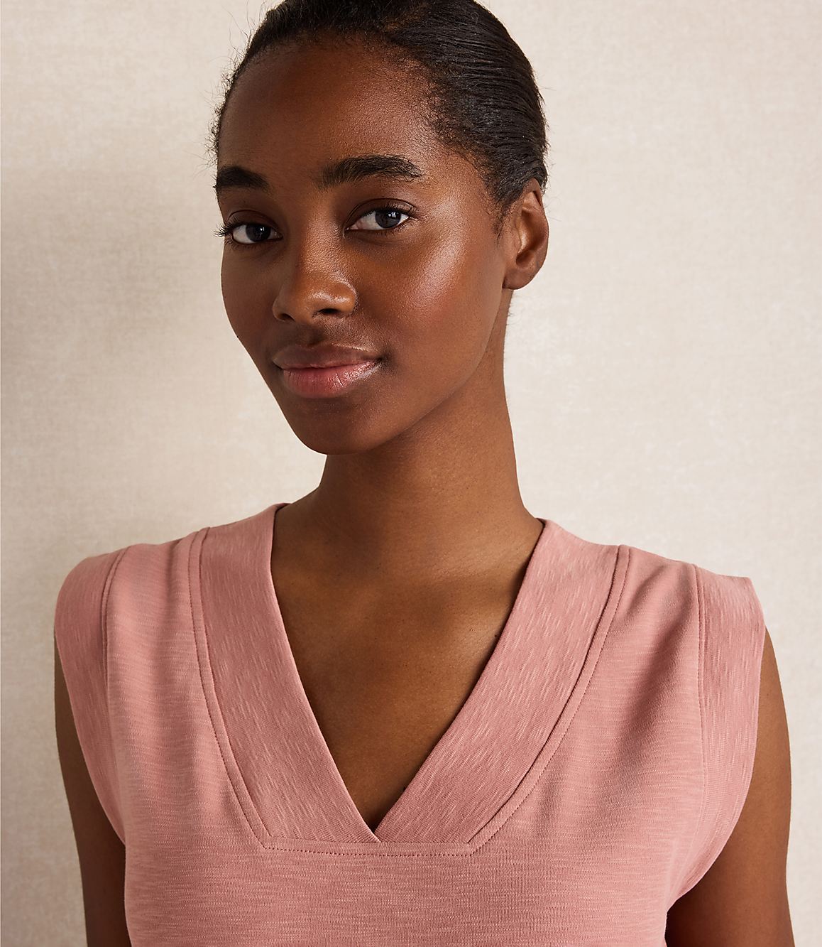 A young woman with dark skin and short hair smiles gently in a pink v-neck top against a textured, light-colored background.