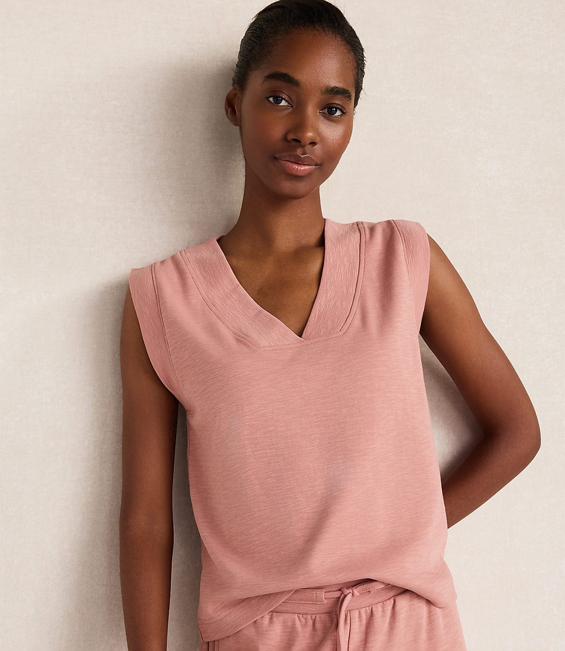 A young woman in a pink v-neck sweater vest stands against a textured, light-colored wall.