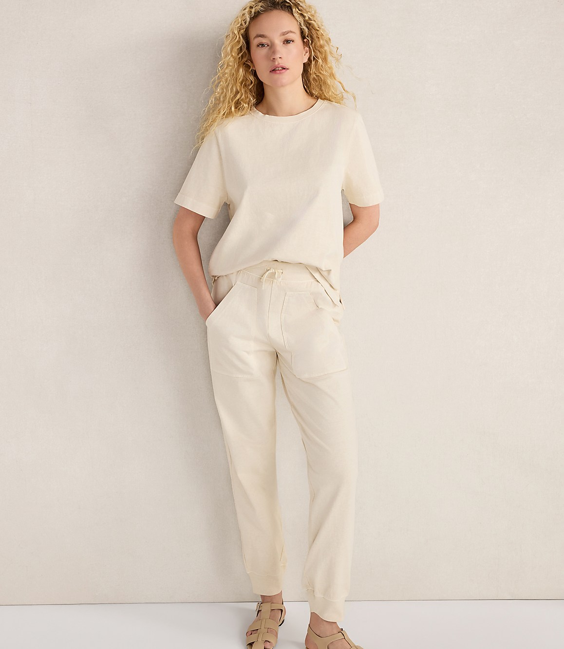A woman in a cream-colored t-shirt and pants stands against a plain, light-colored wall.