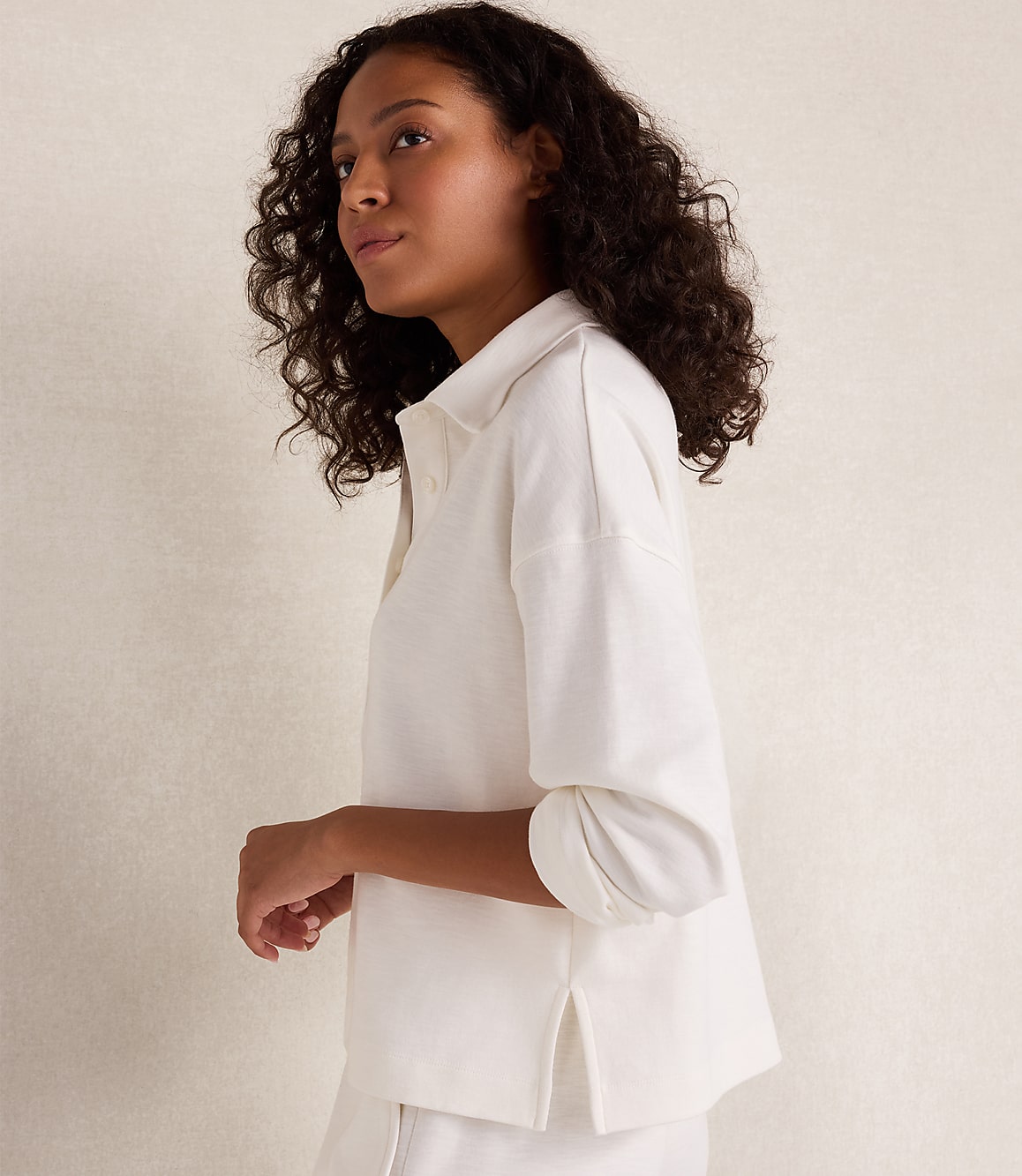 A woman with curly dark hair in a white collared shirt stands against a plain, light-colored background.