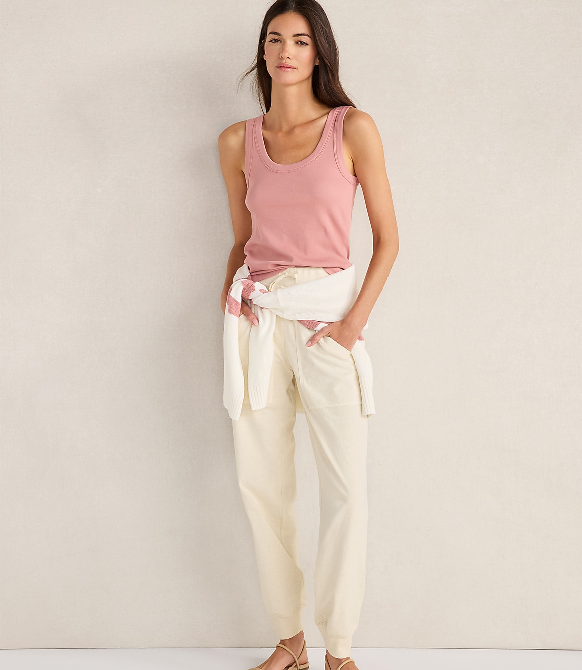A woman in a pink tank top and cream pants stands against a textured, light-colored wall.