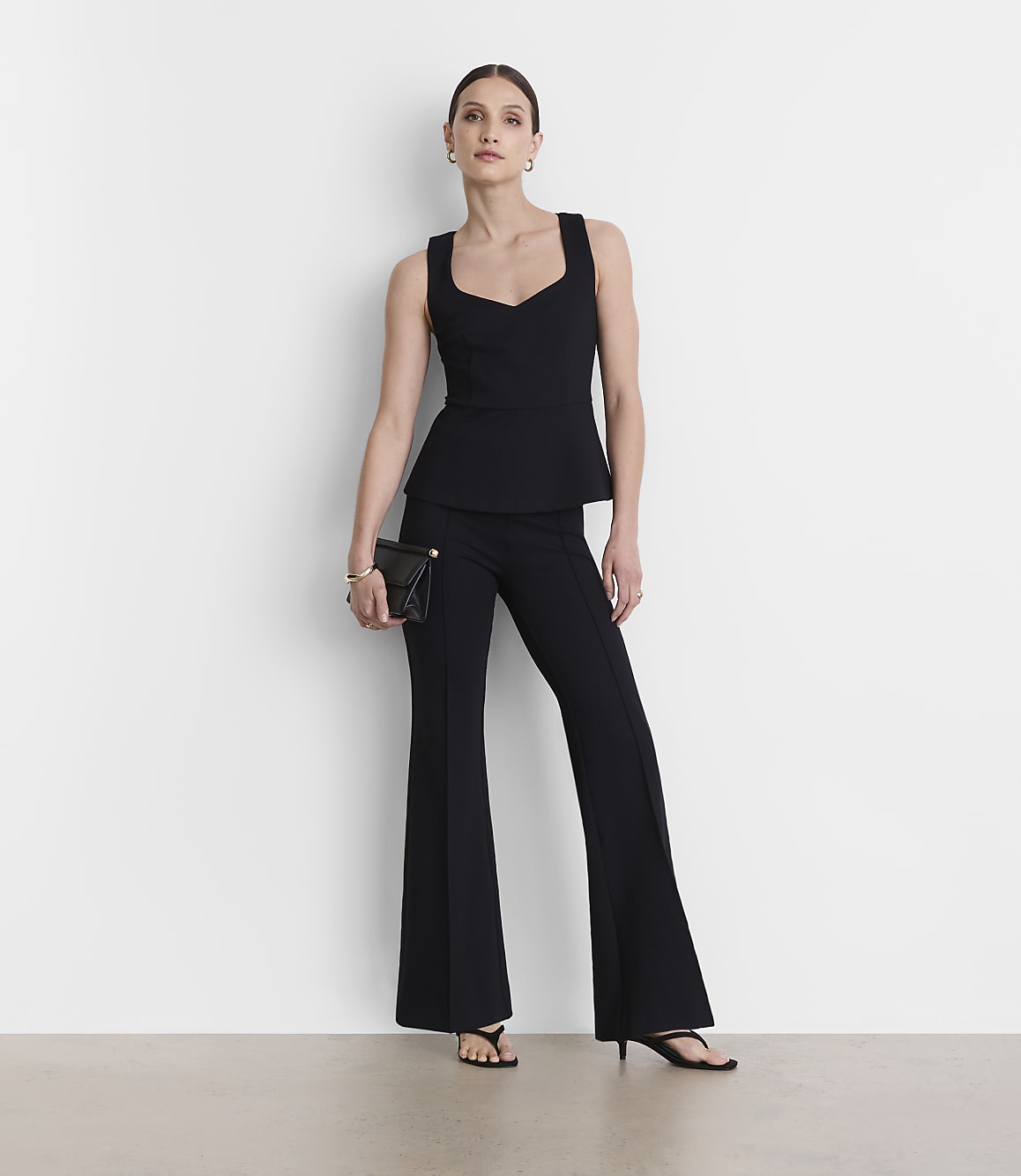 A woman in a black peplum top and flared pants stands against a plain white background, holding a black clutch.