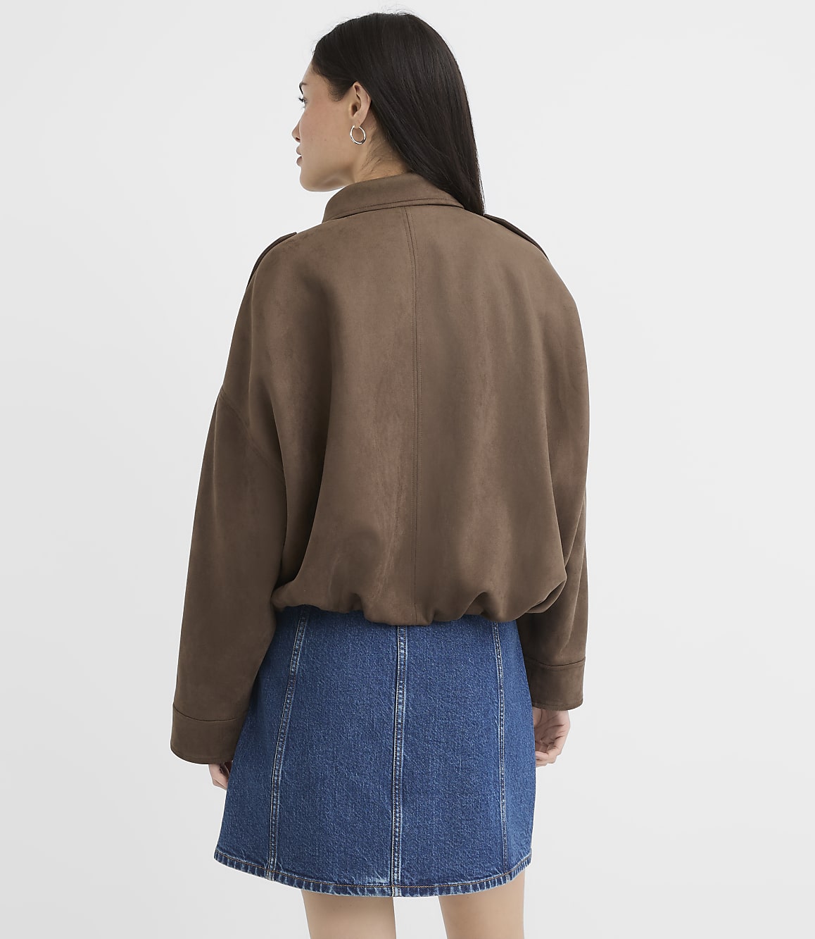A woman in a brown jacket and denim skirt stands against a plain white background.
