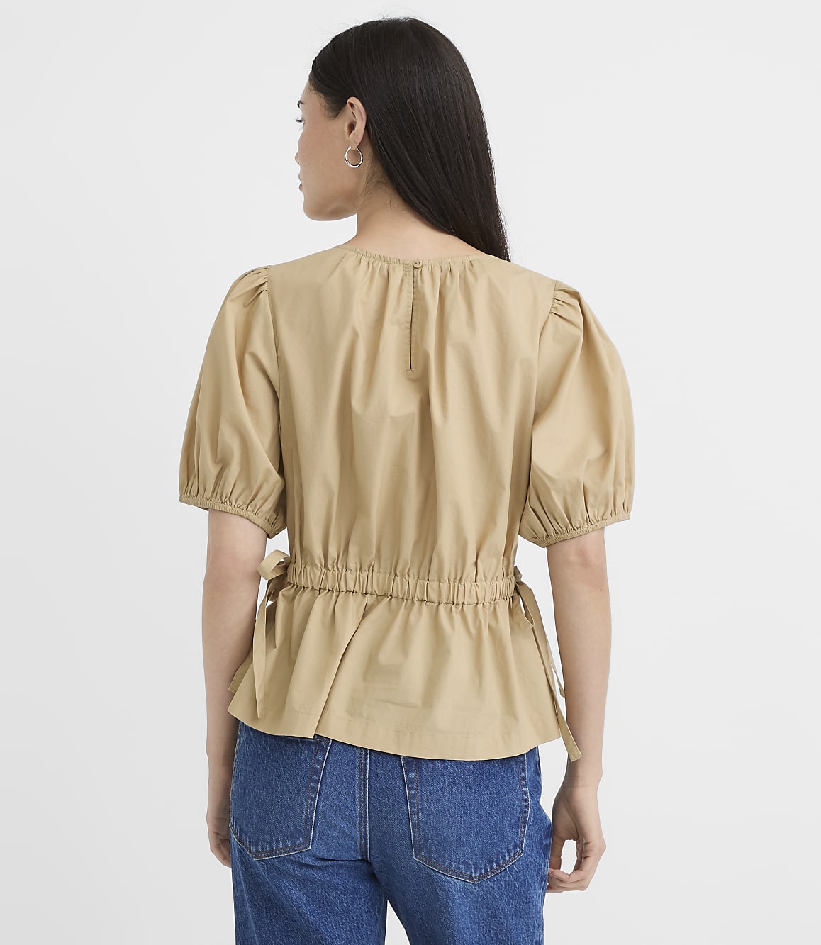 A woman in a tan puff-sleeve top and blue jeans stands against a plain white background.