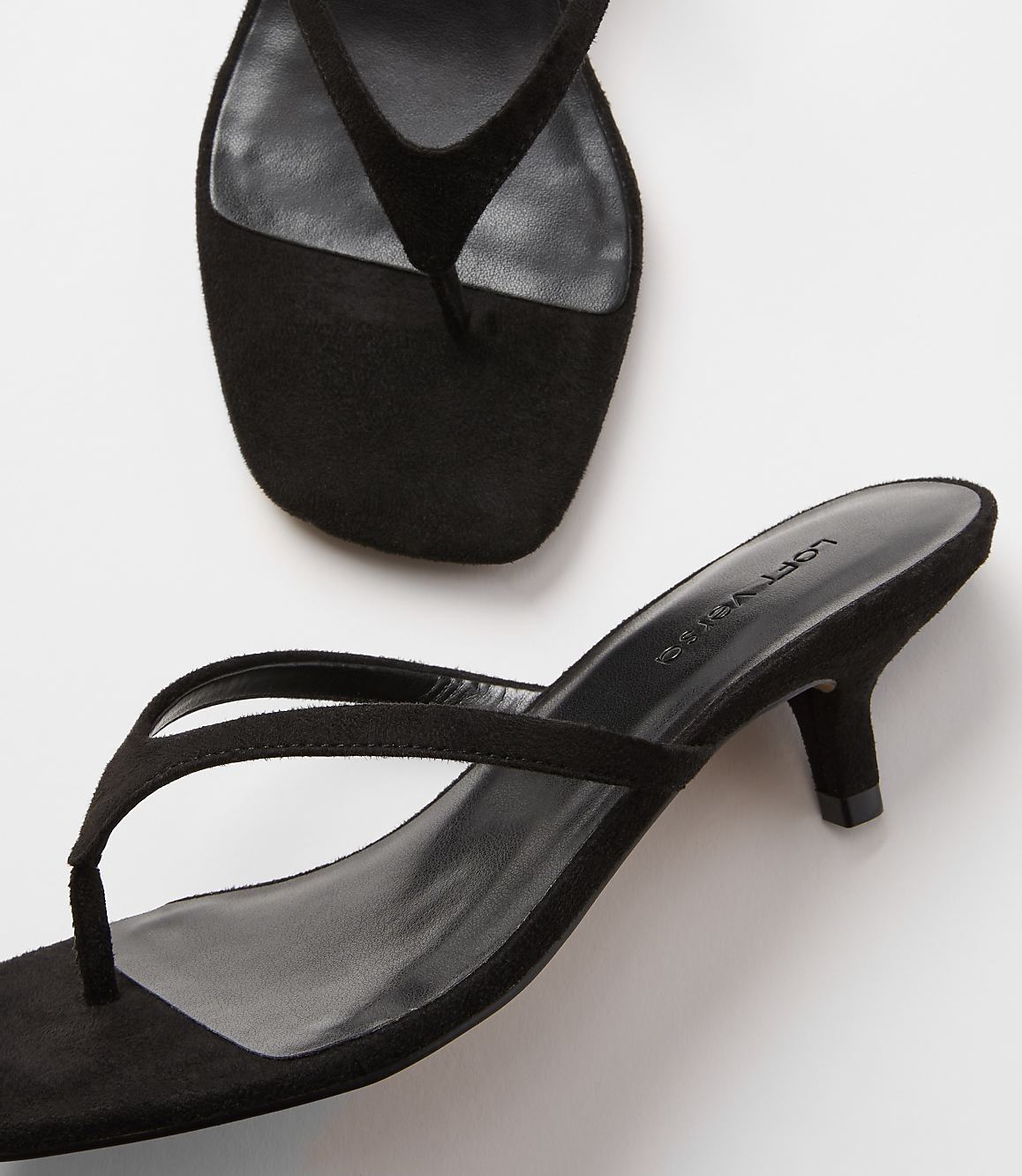 A pair of black suede thong sandals with kitten heels rests on a white surface, with another sandal partially visible in the upper background.