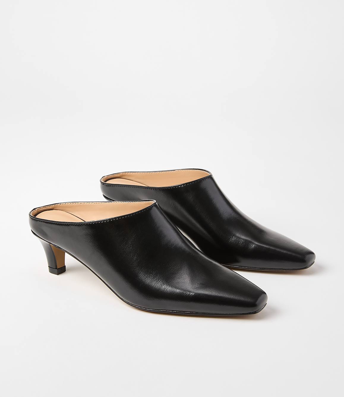 A pair of black leather mules with pointed toes and low heels rests on a plain white background.