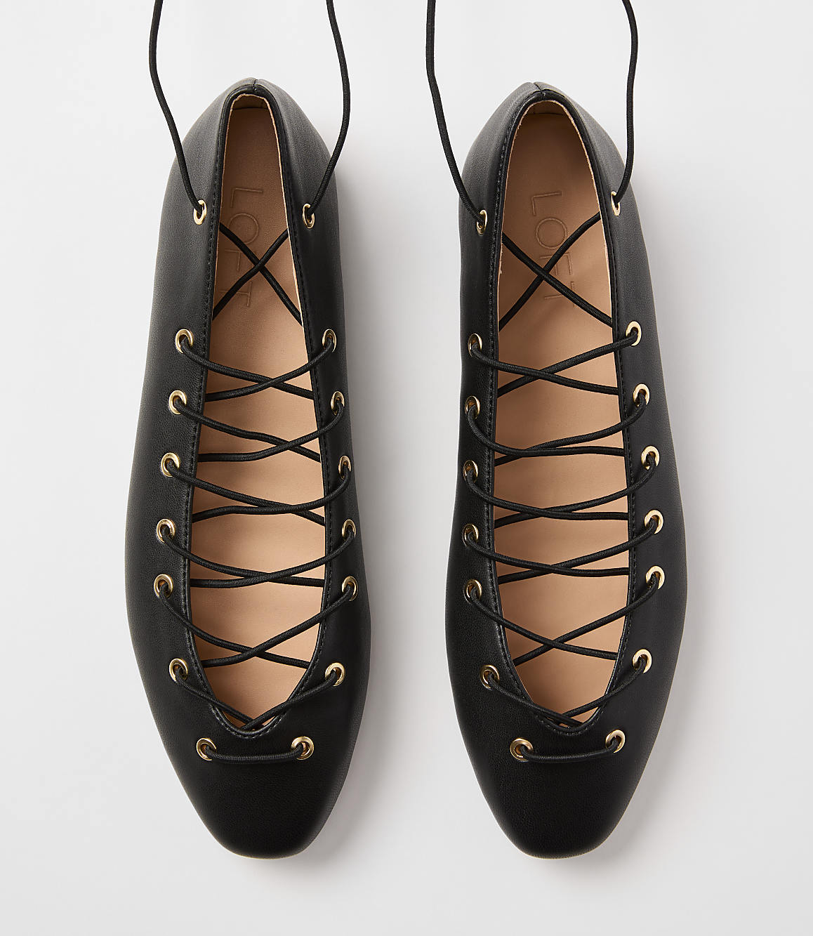 A pair of black lace-up ballet flats with gold eyelets are displayed against a plain white background.