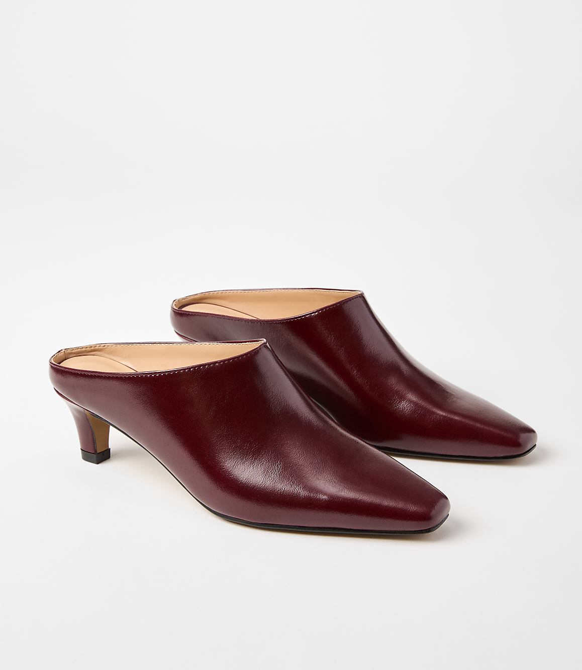 A pair of burgundy leather mules with pointed toes and low heels rests on a plain white background.