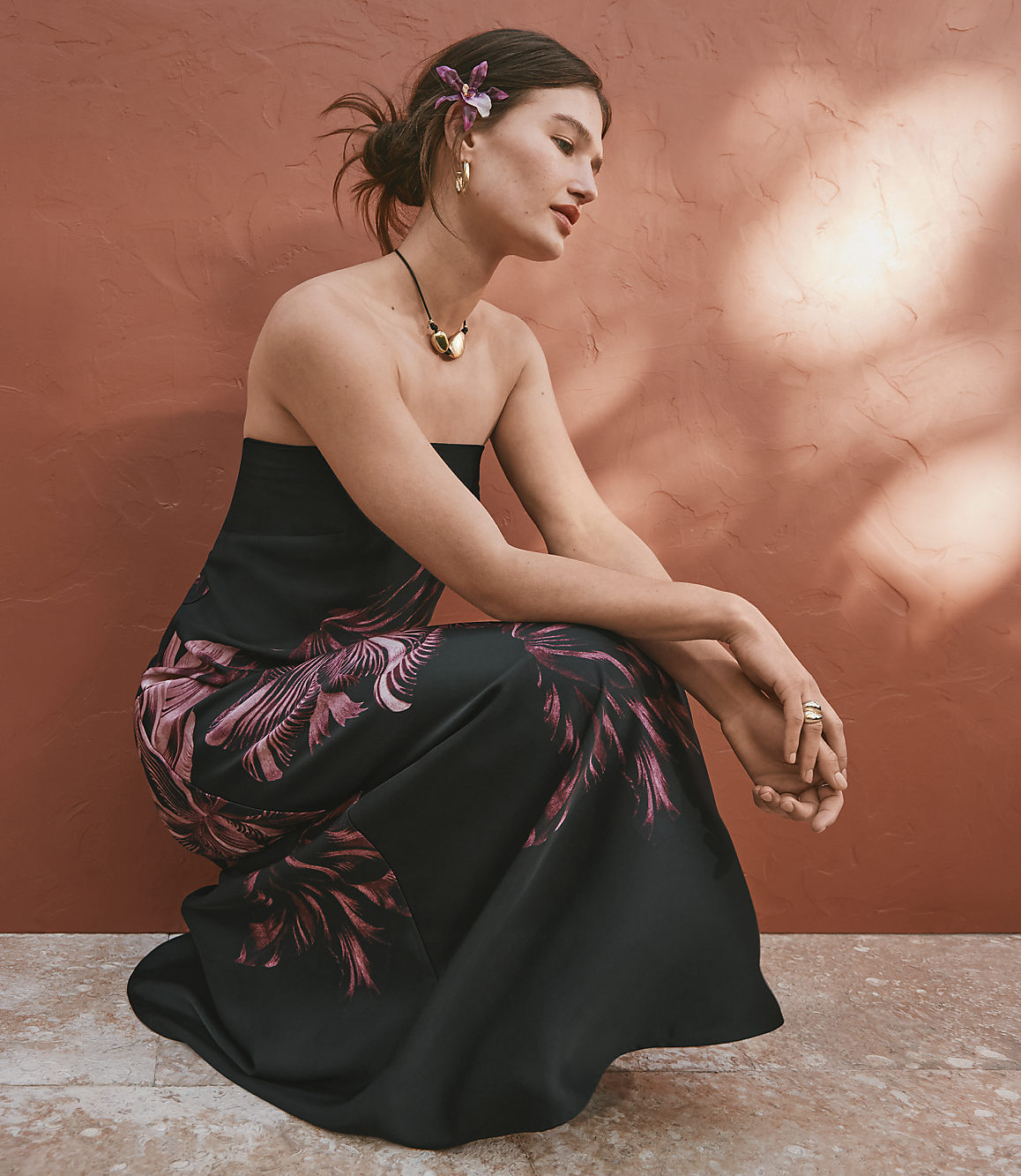 A woman in a strapless floral dress kneels against a terracotta wall with dappled sunlight.