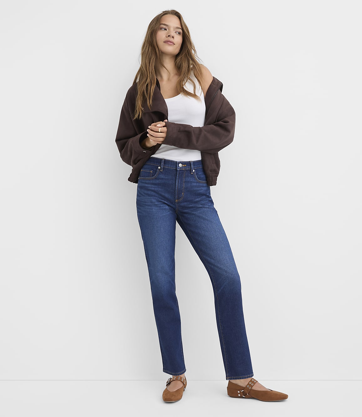 A woman in a white tank top, brown cardigan, and dark wash jeans stands against a plain white background.