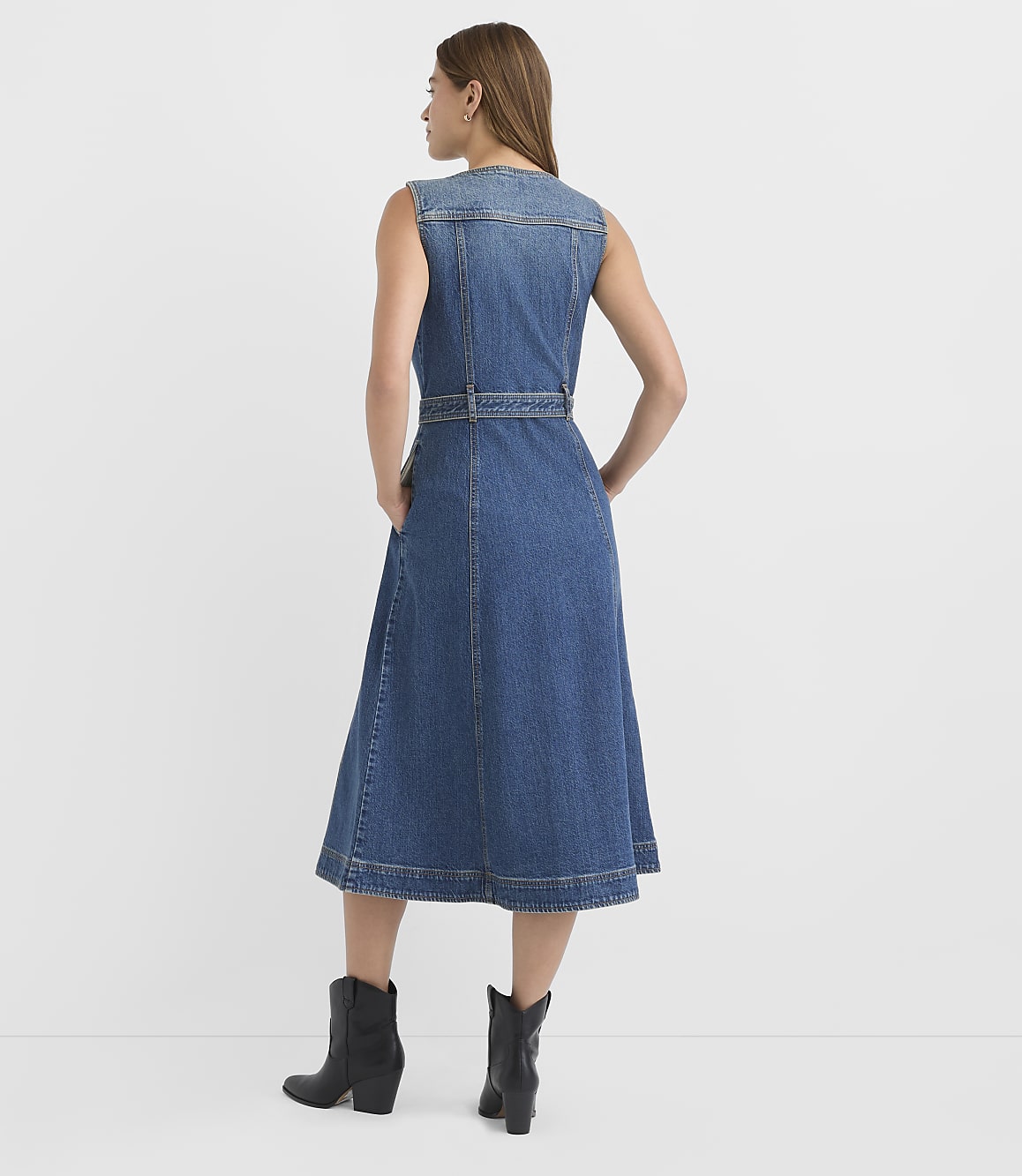A woman in a denim dress and black boots stands with her hands in her pockets against a plain white background.