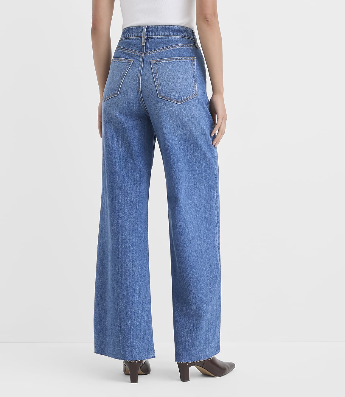 A person wearing wide-leg blue jeans and brown heeled boots stands against a plain white background.