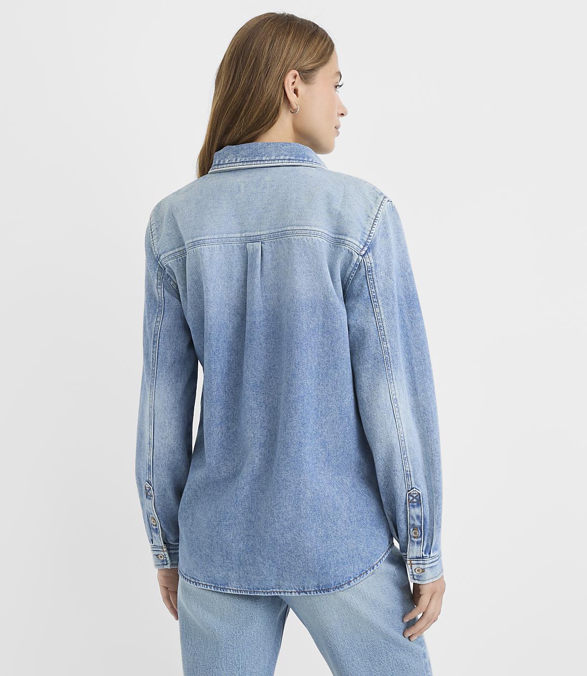 A woman in a light blue denim shirt and jeans stands against a plain white background.