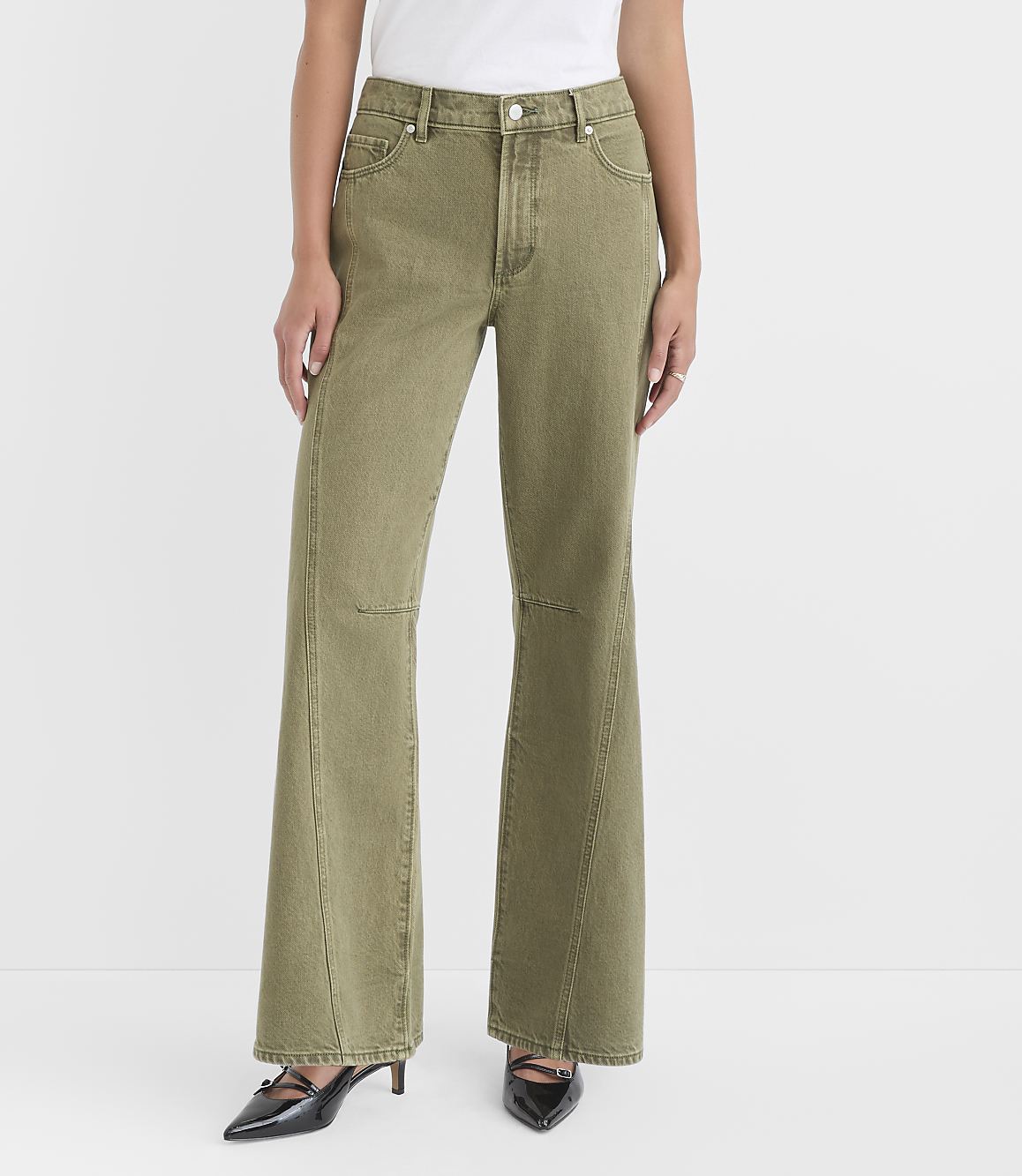 A woman in olive green flared jeans and black heels stands against a plain white background.