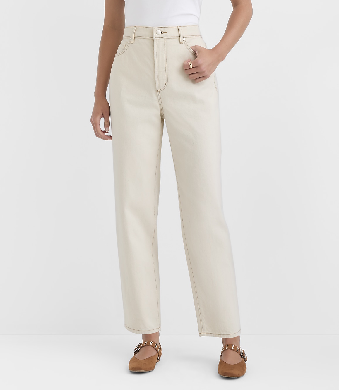 A person stands in the foreground wearing cream-colored jeans and brown flats against a plain white background.