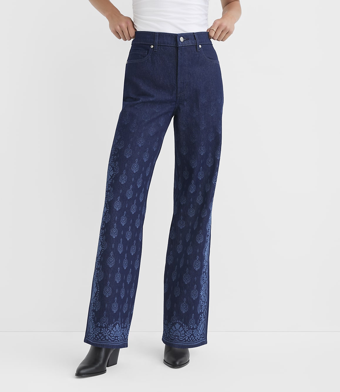 A person stands in dark wash flared jeans with a blue paisley pattern on the lower legs and a plain white background.