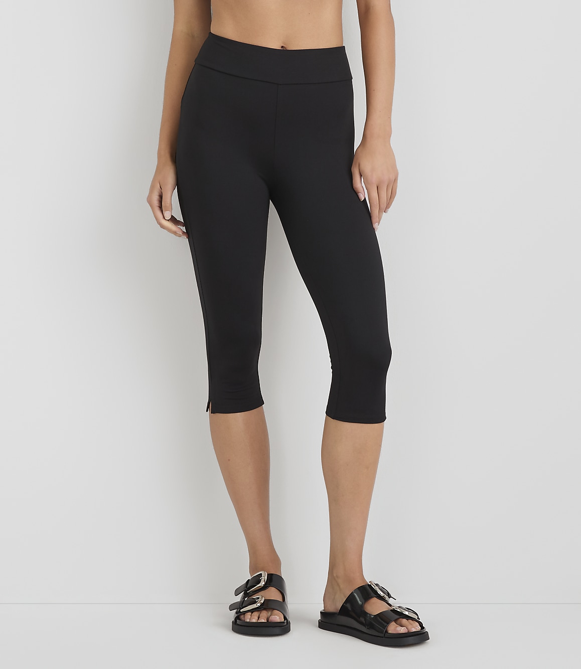 A person wearing black capri leggings and black sandals stands against a plain white background.