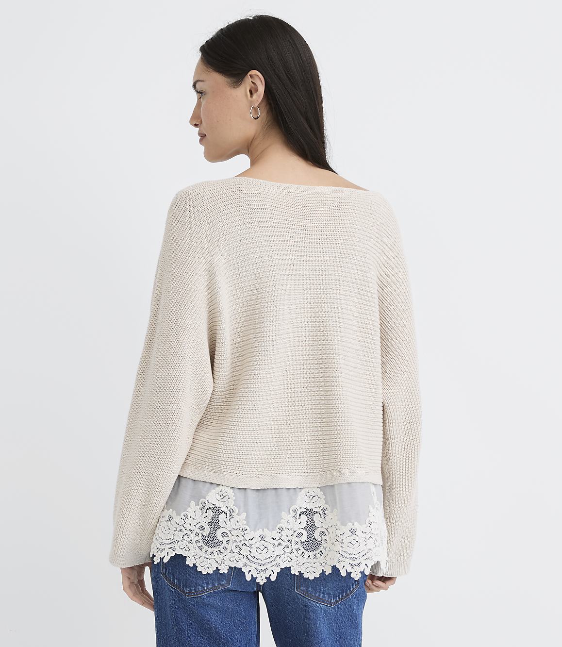 A woman in a cream-colored sweater with a lace hem stands against a plain white background.