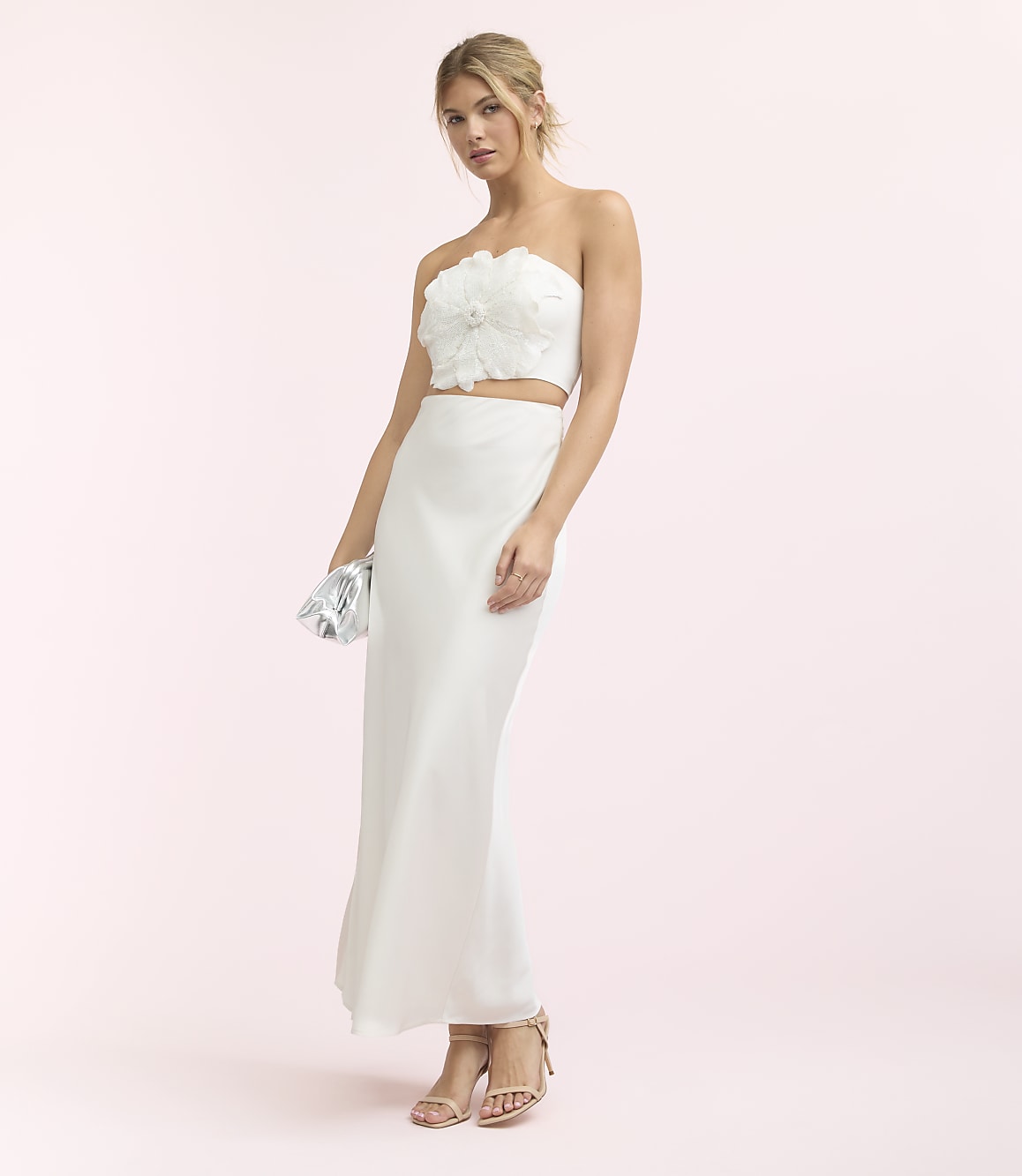 A woman in a white strapless top and skirt stands against a soft pink background, holding a silver clutch.