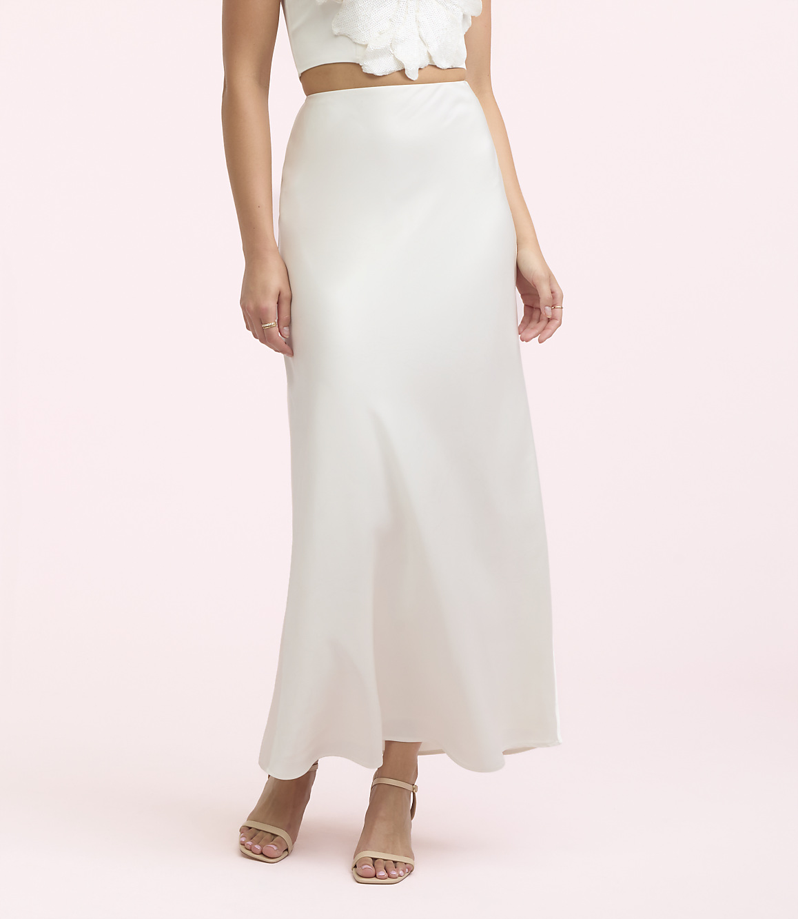 A woman in a white satin skirt and sandals stands against a soft pink background.