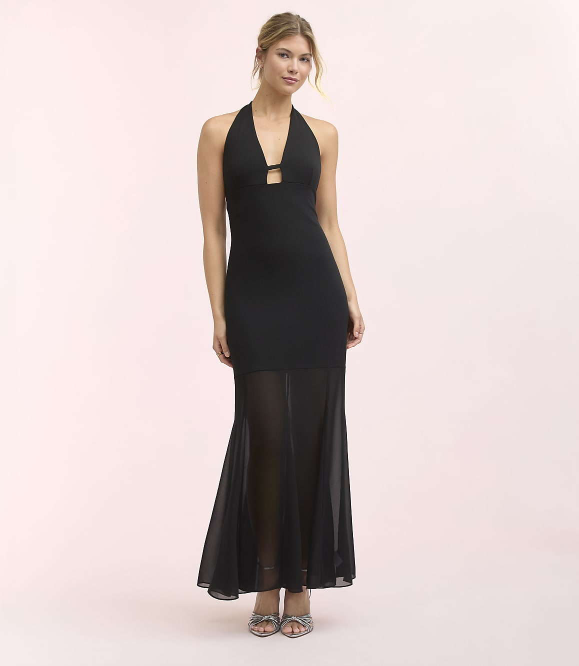 A woman in a black halter dress with a sheer skirt stands against a soft pink background.