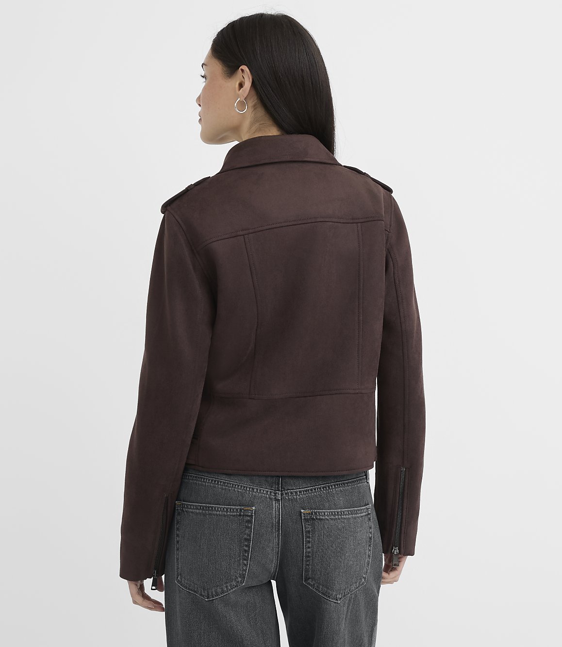 A woman in a brown jacket and jeans stands against a plain white background.