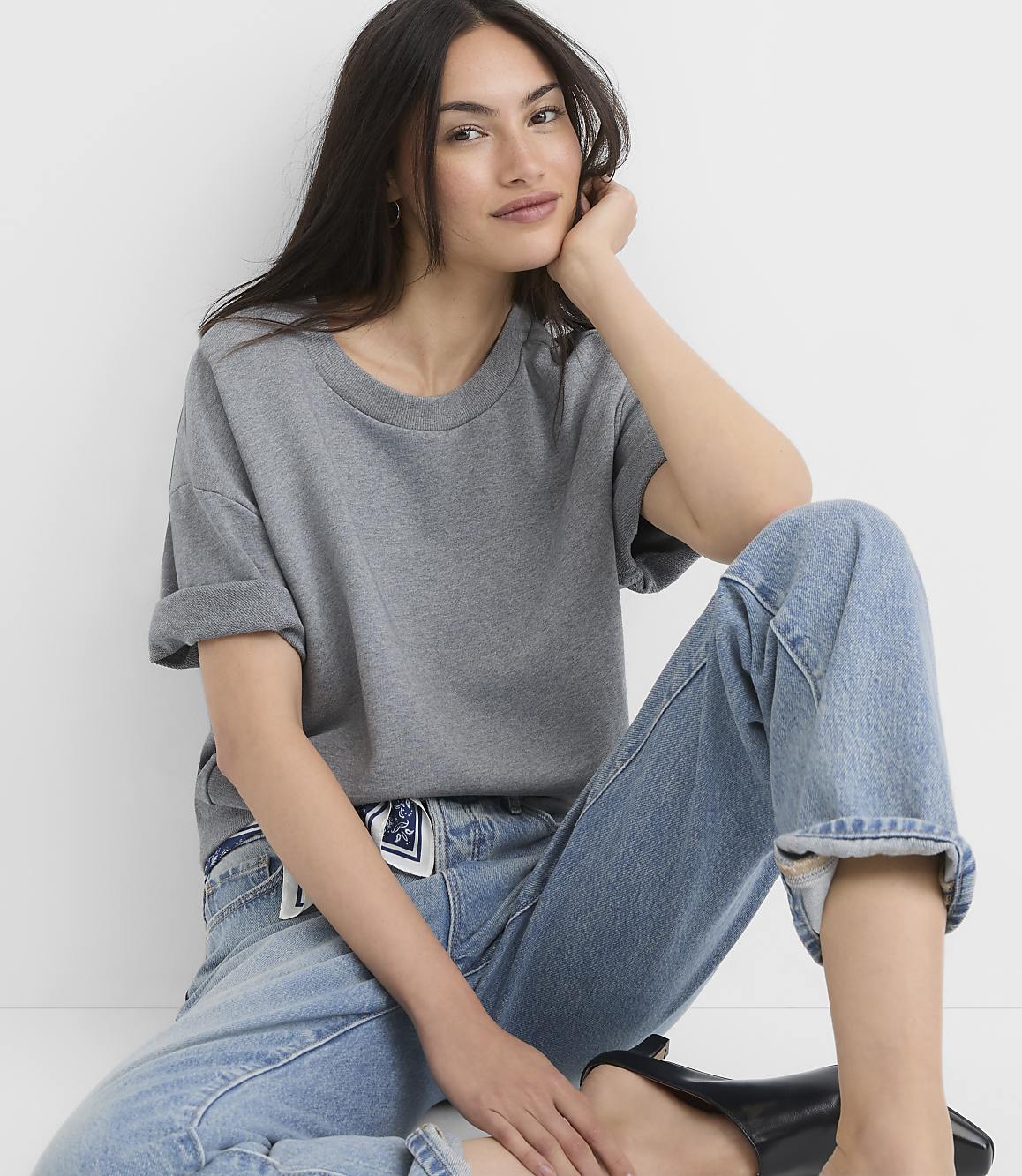 A young woman in a grey t-shirt and blue jeans sits against a plain white background.
