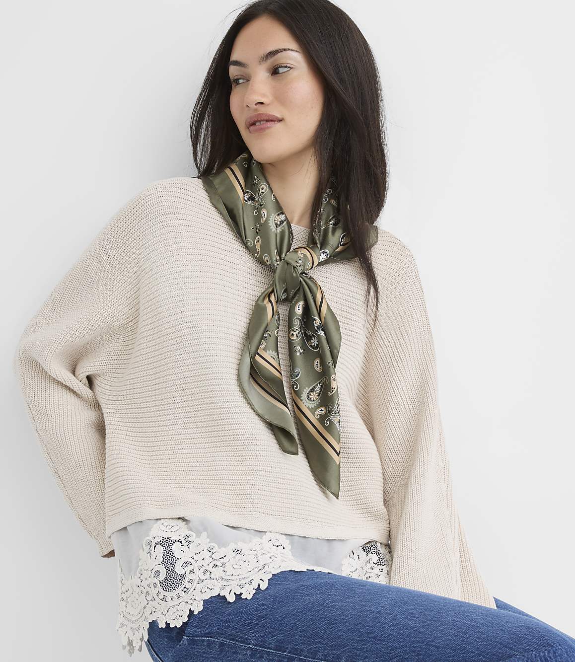 A woman in a cream sweater and jeans, accessorized with a green patterned scarf, poses against a plain white background.