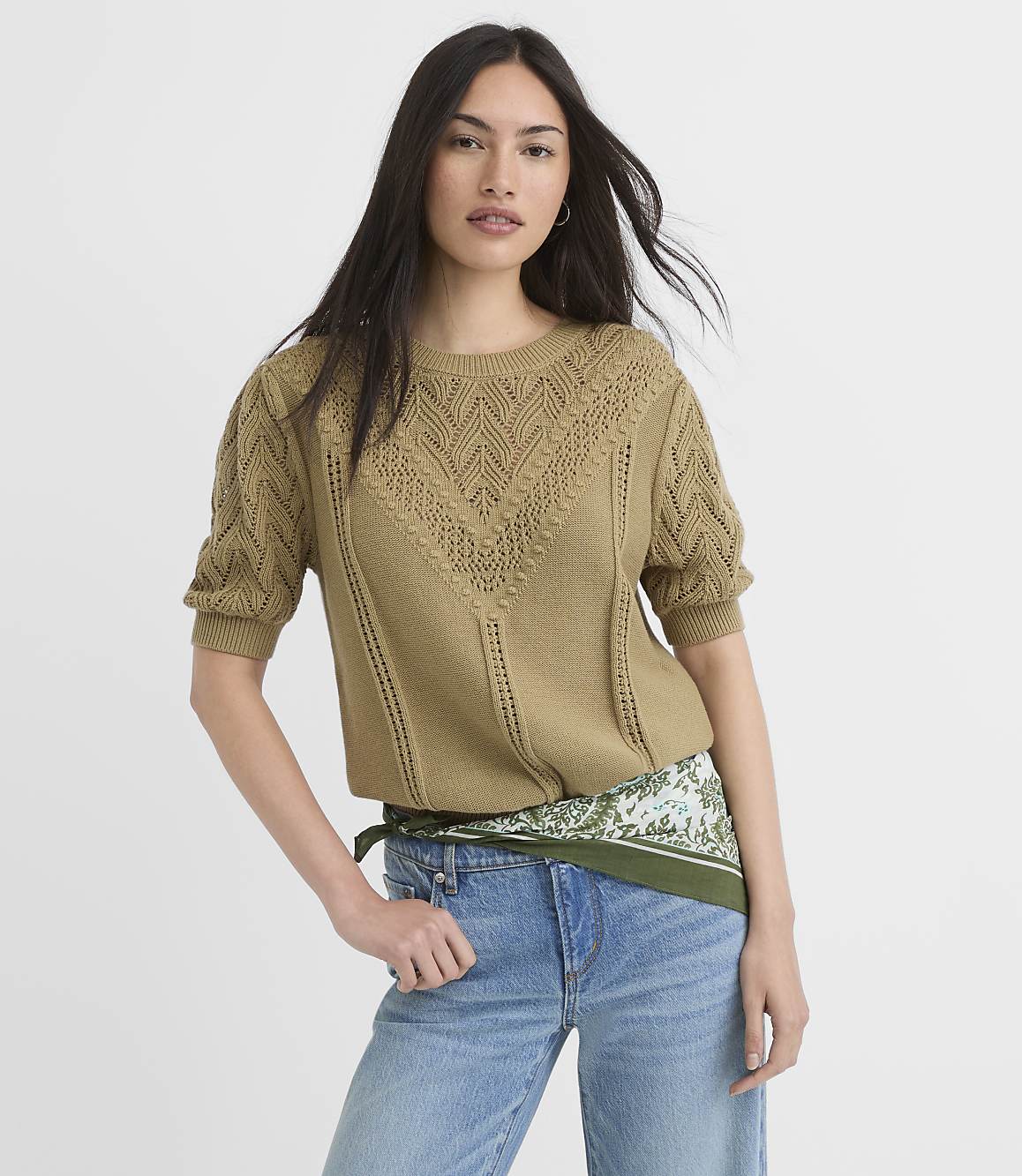 A woman in a textured olive green sweater and light wash jeans stands against a plain white background.