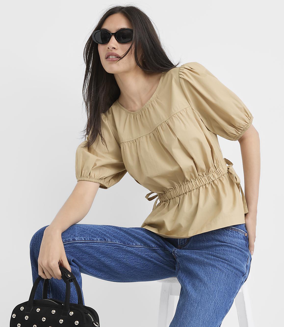 A woman in a tan puff-sleeve top and blue jeans, wearing sunglasses and holding a studded black bag, poses against a plain white background.