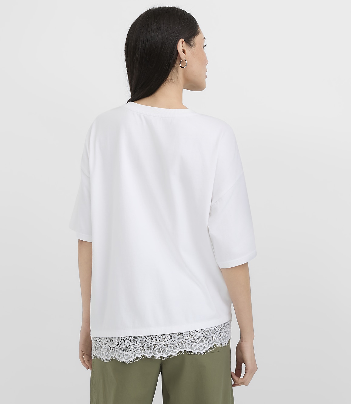 A woman in a white t-shirt with lace trim stands against a plain white background.