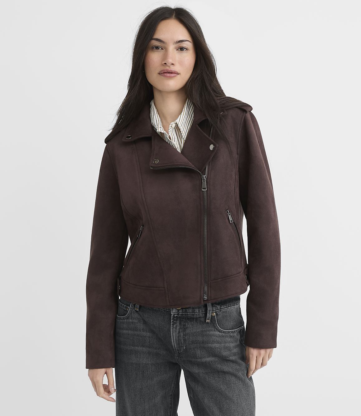 A woman in a brown suede moto jacket and jeans stands against a plain, light brown background.