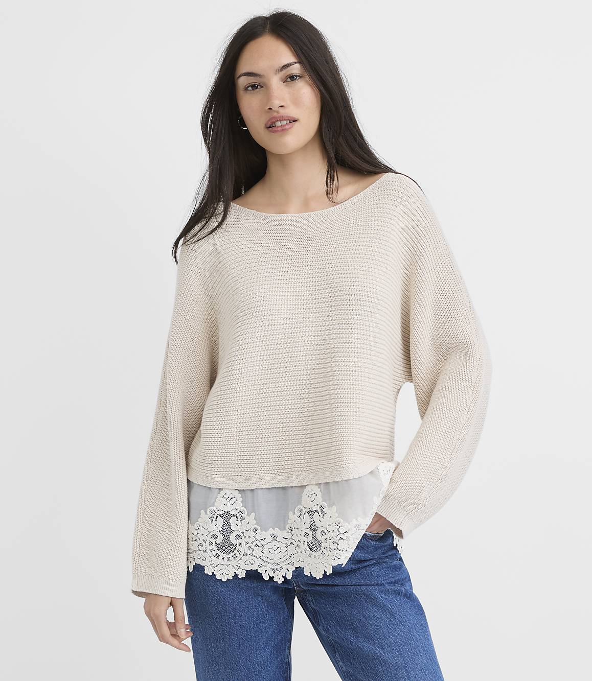 A woman in a cream sweater and blue jeans stands against a plain white background.