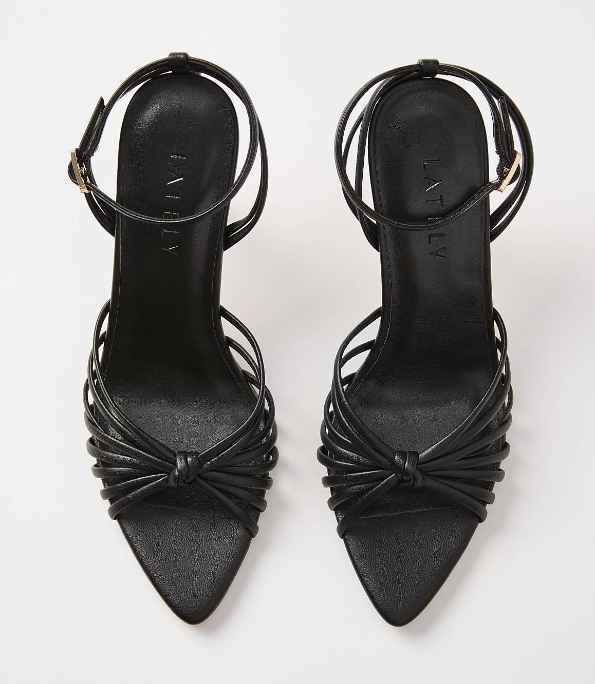 A pair of black strappy high-heeled sandals with knotted details in the foreground are set against a plain white background.