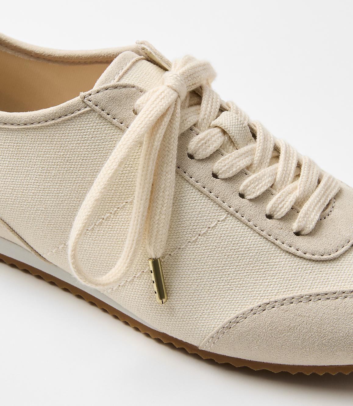 A close-up of a cream-colored canvas sneaker with beige suede accents and laces is set against a plain white background.