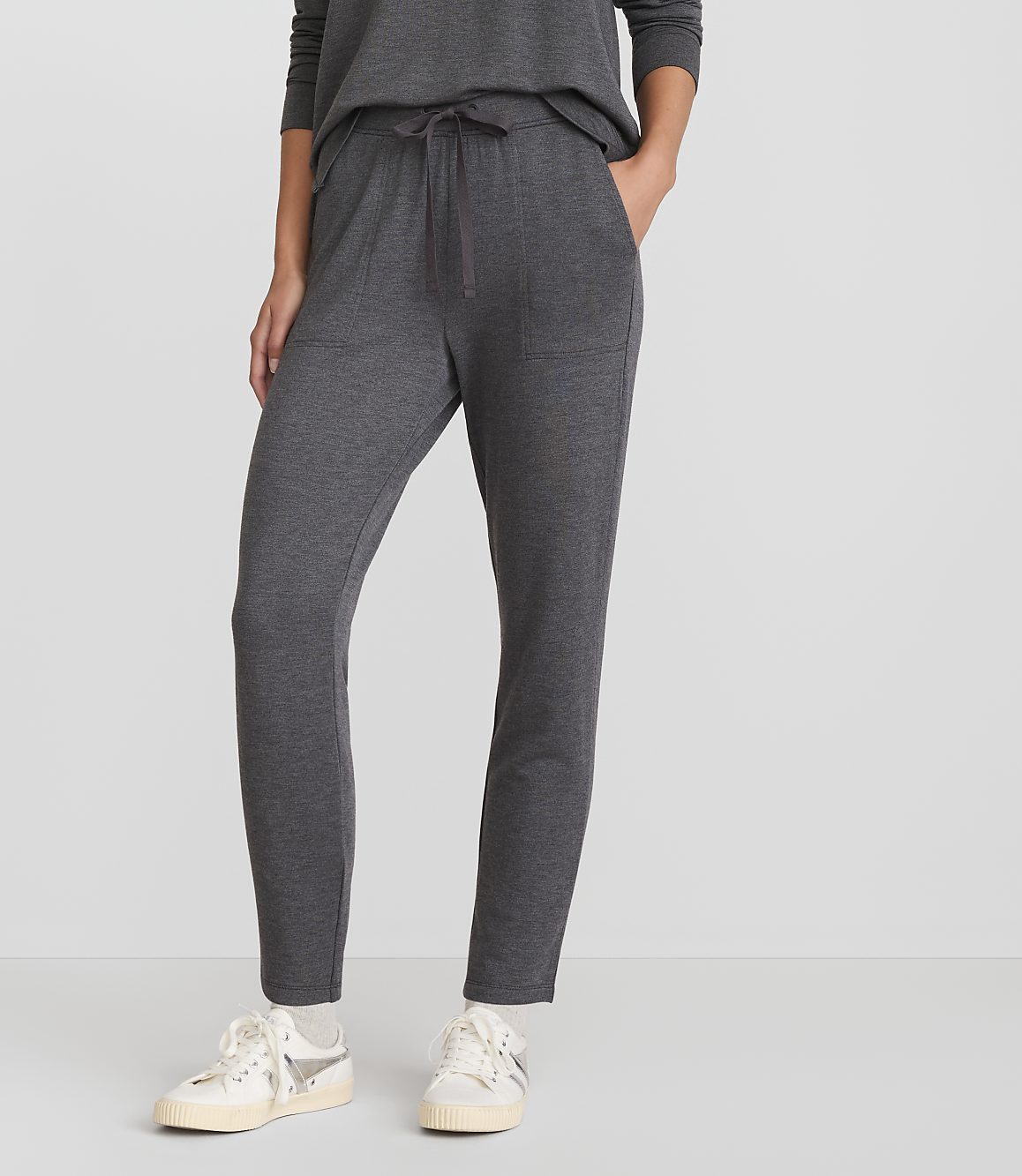 A person wearing dark gray sweatpants and white sneakers stands against a plain white background.