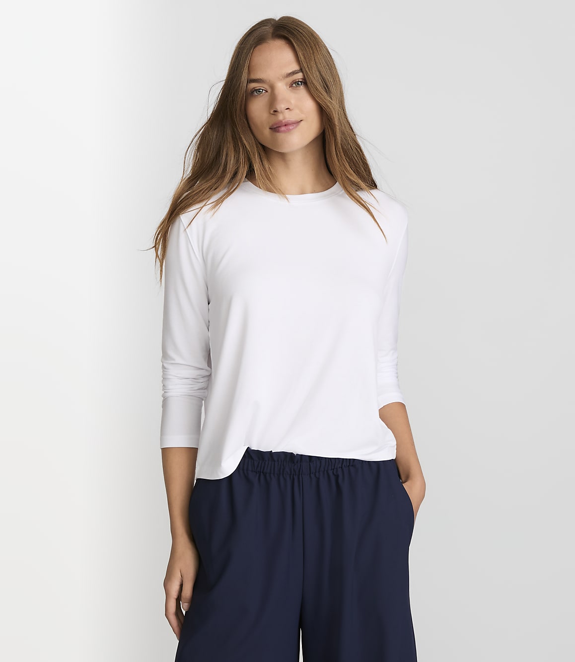 A woman in a white long-sleeved shirt and navy pants stands against a plain white background.