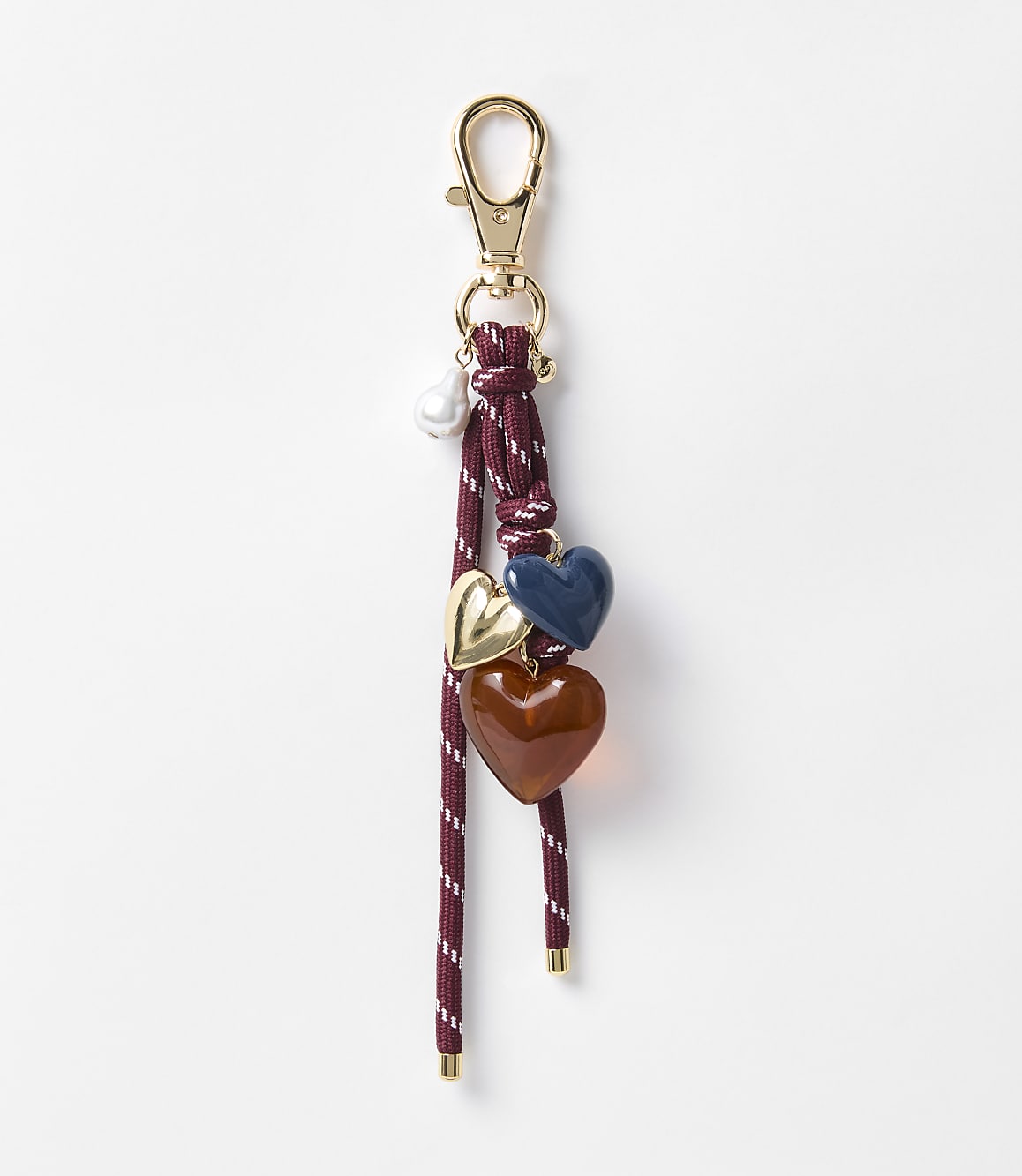 A burgundy and white striped cord keychain with gold hardware dangles three heart charms&mdash;one gold, one navy blue, and one amber&mdash;against a plain white background.