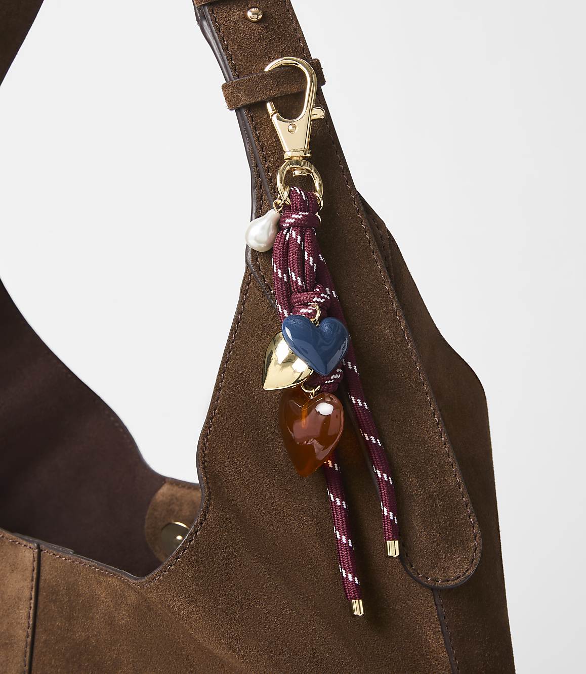A brown suede bag with a decorative charm featuring hearts and a pearl hangs in the foreground, against a plain white background.