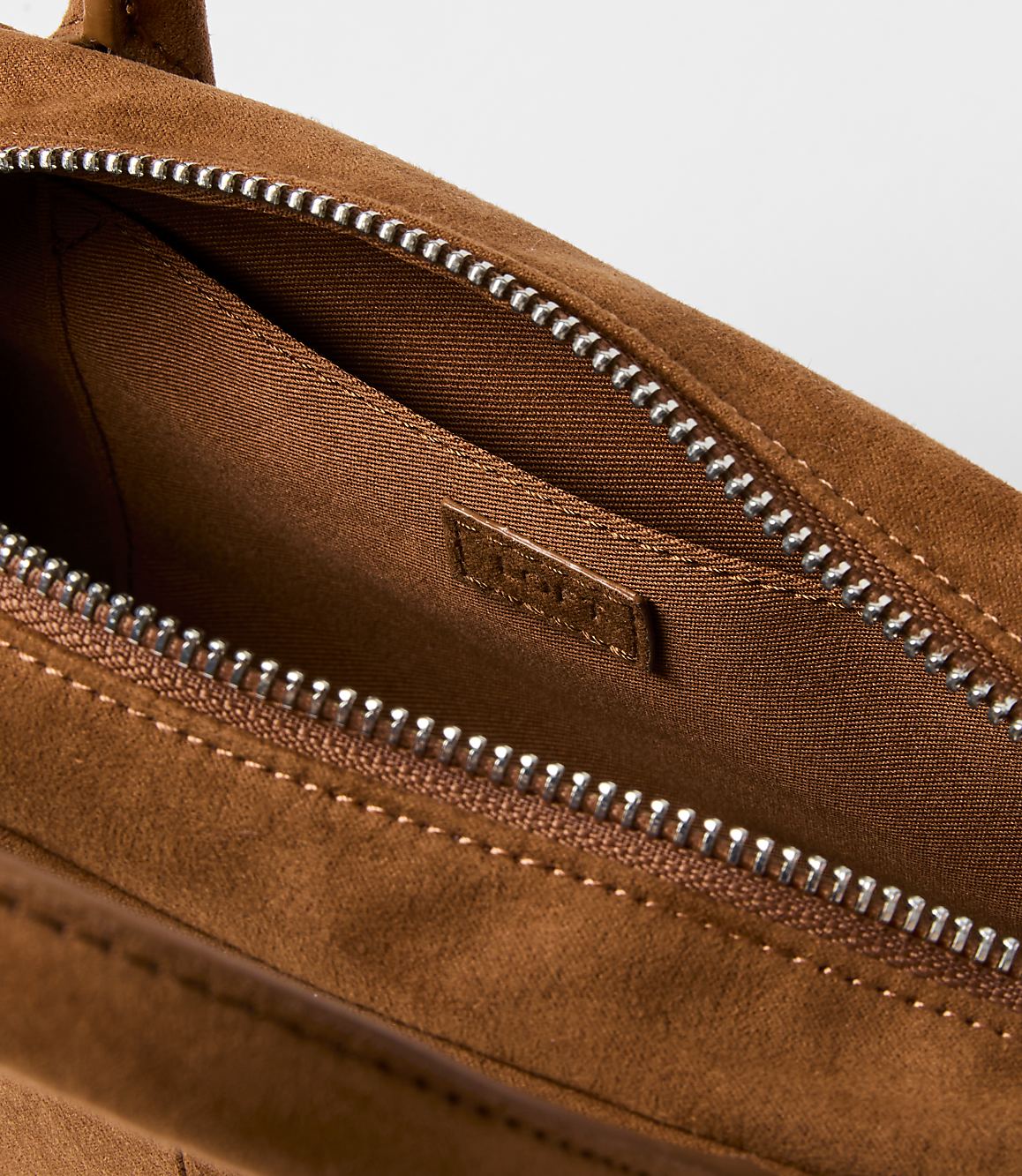 A close-up of an open brown suede handbag with a silver zipper in the foreground, set against a plain white background.