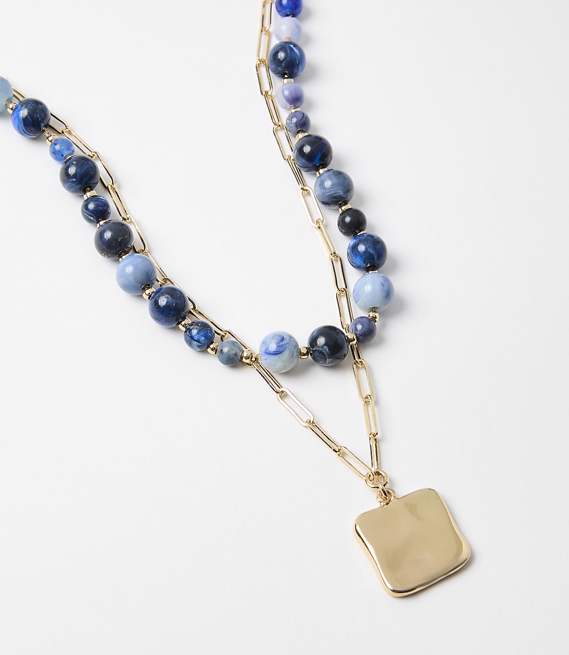 A layered necklace with blue beads and a gold square pendant rests on a plain white background.