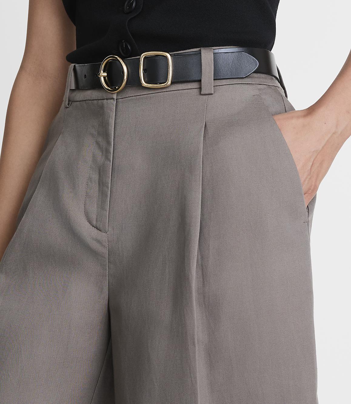 A person's hands are in the pockets of taupe-colored, pleated shorts with a black belt, against a plain white background.