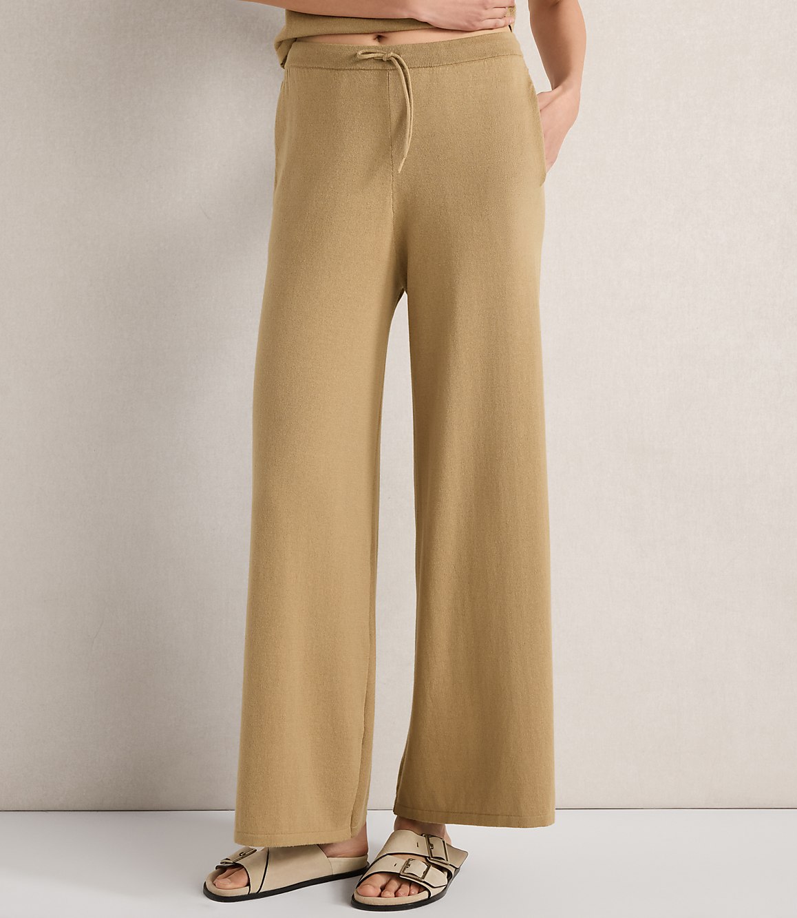 A person stands in tan wide-leg pants and sandals against a textured off-white background.