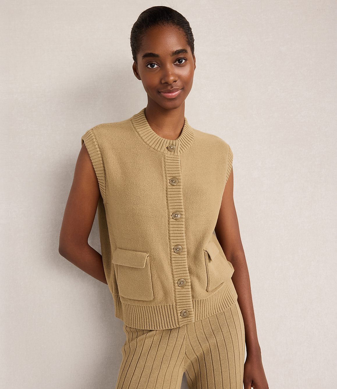 A woman in a tan knit vest and matching pants stands against a plain, light-colored background.