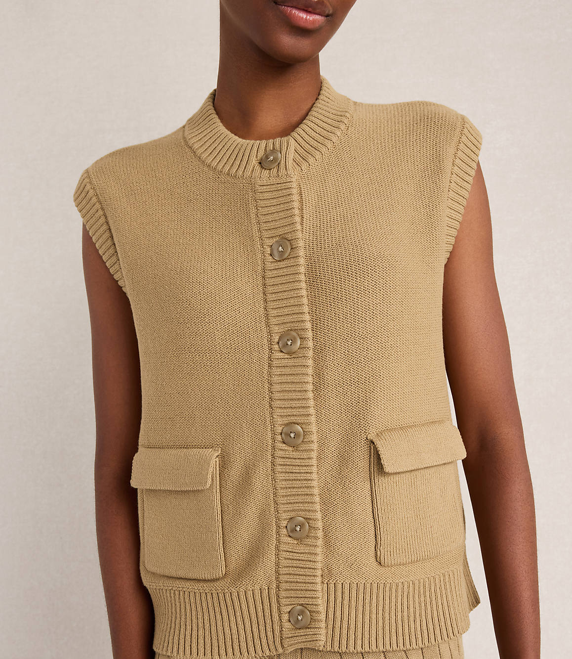 A close-up of a person wearing a tan knit vest with buttons and pockets is set against a plain, light-colored background.